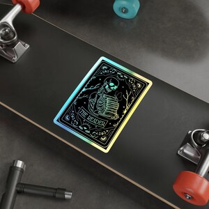 Holographic tarot card sticker featuring a skeleton holding a stack of books with a butterfly atop, surrounded by celestial stars and tree branches. Perfect for laptops, journals, water bottles, and scrapbooks. Ideal gift for readers and book lovers.