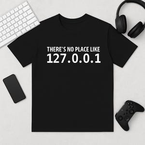 Peut inclure: T-shirt noir avec l'inscription blanche "THERE'S NO PLACE LIKE 127.0.0.1". Le t-shirt est entouré d'un clavier, d'un casque, d'un smartphone et d'une manette de jeu.