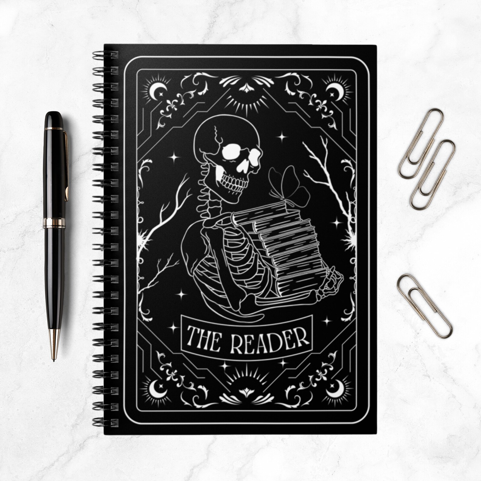 A black notebook with a tarot card drawing on it in white sitting on a marble background. The tarot card drawing has a white outline with a skeleton holding a pile of books in it. It says "The Reader" at the bottom, along with some other various designs.