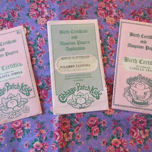 May include: Three Cabbage Patch Kids birth certificates and adoption papers on a floral patterned background. The certificates are pink and green, with text including names and the Cabbage Patch Kids logo.