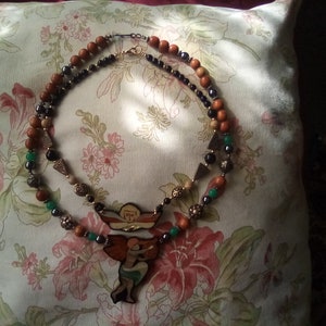 May include: A multi-strand necklace with brown wooden beads, black beads, green beads, and a gold pendant featuring two angels. The pendant is painted with colorful details.