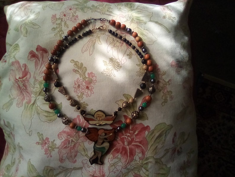 May include: A multi-strand necklace with brown wooden beads, black beads, green beads, and a gold pendant featuring two angels. The pendant is painted with colorful details.