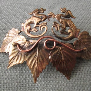 May include: A vintage metal brooch featuring two stylized dragons facing each other. The dragons are gold-toned, perched above gold and copper-colored leaves. A copper wire adds a decorative element to the design.