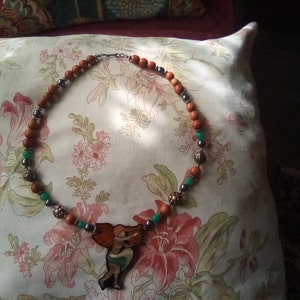 May include: A brown and green beaded necklace with a wooden pendant featuring a woman holding a ball. The pendant is painted with orange, green, and brown colors.