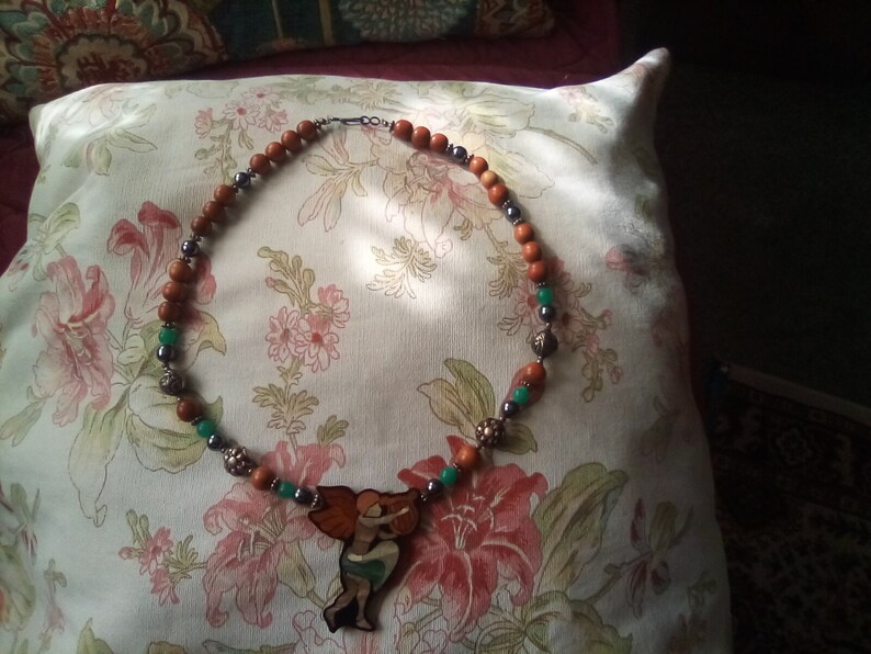 May include: A brown and green beaded necklace with a wooden pendant featuring a woman holding a ball. The pendant is painted with orange, green, and brown colors.