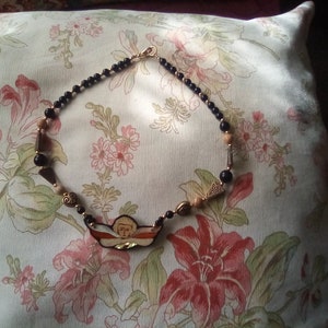 May include: A black and gold beaded necklace with a painted angel pendant. The pendant is a light brown, orange, and white color with a gold halo. The necklace is on a floral patterned pillow.