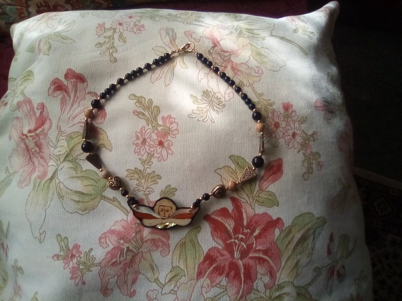 May include: A black and gold beaded necklace with a painted angel pendant. The pendant is a light brown, orange, and white color with a gold halo. The necklace is on a floral patterned pillow.