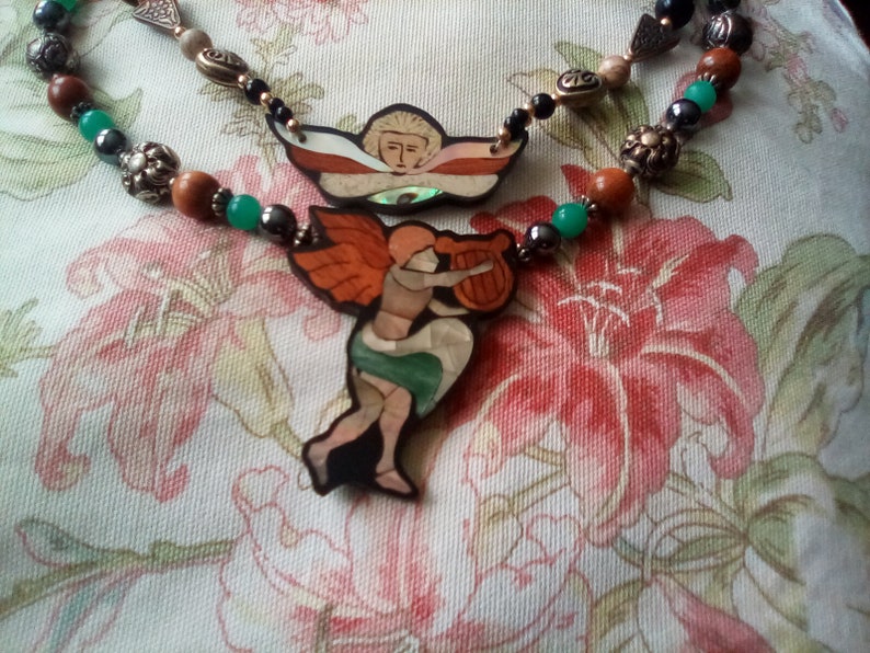 May include: A necklace with green, brown, and black beads and two pendants. The pendants are made of wood and feature painted images of angels.