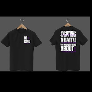 May include: Two black t-shirts hanging on wooden hangers. The first shirt has the words "BE KIND" in white and purple. The second shirt has the text "EVERYONE YOU MEET IS FIGHTING A BATTLE YOU KNOW NOTHING ABOUT" in white and purple.