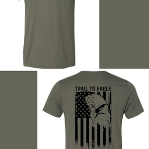 Könnte beinhalten: Olivgrünes T-Shirt mit einem schwarzen Pfadfinder-Emblem auf der Vorderseite. Die Rückseite des Shirts zeigt ein amerikanisches Flaggen-Design mit einem Adler und dem Text "TRAIL TO EAGLE".