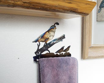 May include: A metal wall-mounted towel rack shaped like a quail with its chicks on a branch. The rack is painted in shades of blue and brown. A purple towel is hanging from the rack.