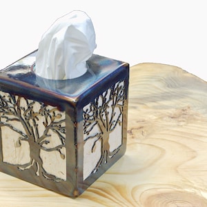 以下が含まれることがあります： 木製のデザインが施された正方形のセラミック製のティッシュボックスホルダー。ホルダーはメタリックブルーの釉薬で装飾されており、上から白いティッシュがのぞいています。