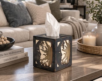 Puede incluir: Un dispensador de pañuelos cuadrado de metal negro envejecido con diseños calados dorados. Está sobre una mesa de centro de madera, con libros y un cuenco decorativo. Se ven pañuelos blancos.