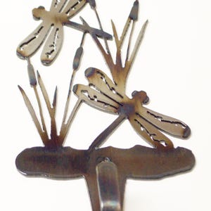 May include: Metal wall hook with a dragonfly design. The hook is shaped like two dragonflies perched on reeds. The hook is a dark brown color with a rustic finish.