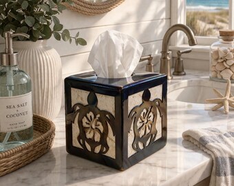 May include: A square tissue box with a dark blue border and a cream-coloured centre. The centre features a cut-out sea turtle design. The box is on a marble countertop next to a bottle of hand soap and a decorative vase.