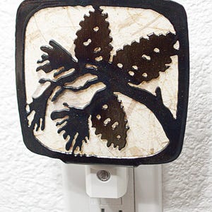 May include: A black metal nightlight with a cutout design of a pine tree with three pine cones. The nightlight is plugged into a standard electrical outlet.