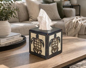 May include: A square tissue box with a dark blue frame and off-white panels featuring a brown turtle design. The box is filled with white tissues and sits on a wooden coffee table. The box is a decorative home accessory.