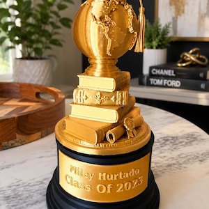 May include: A gold-colored trophy featuring a stack of books, a rolled diploma, and a globe topped with a graduation cap. The black circular base displays the gold-colored text: Miley Hurtado Class Of 2023. The trophy has a metallic finish.