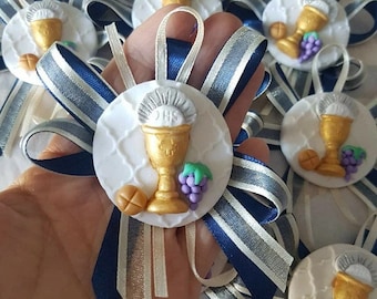 May include: A collection of religious-themed favors. Each favor features a white circular base with a gold chalice, grapes, and a small bread roll. The favors are adorned with blue, silver, and white ribbons.