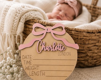 May include: A circular wooden birth announcement sign featuring a light pink cutout bow at the top. The name Charlotte is written in pink script, with the text LONDON and the labels DATE, TIME, WEIGHT, and LENGTH printed in black below.