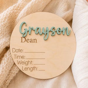 May include: A circular light-colored wooden plaque featuring the name Grayson in raised teal script lettering and the name Dean in brown text. Below, the words Date, Time, Weight, and Length are printed with blank lines for custom information.