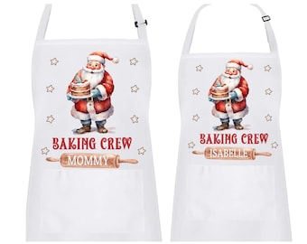 May include: Two white aprons with adjustable neck straps. Each apron features a watercolor illustration of Santa Claus holding a cake, surrounded by stars. The text "BAKING CREW" and a rolling pin graphic are below the Santa image. One apron says "MOMMY" and the other says "ISABELLE".