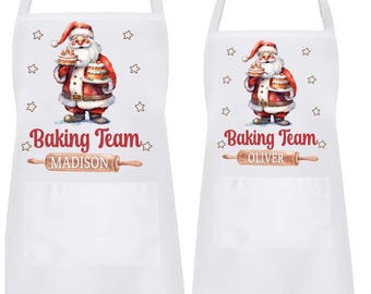 May include: White aprons with a Santa Claus graphic holding a cake, the words "Baking Team" and a name below a rolling pin. The aprons have a pocket and adjustable neck strap. One apron says "Madison" and the other says "Oliver".