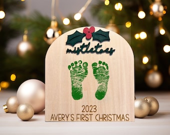 May include: A wooden Christmas ornament with a green mistletoe design, two green footprints, and the text "2023 AVERY'S FIRST CHRISTMAS".