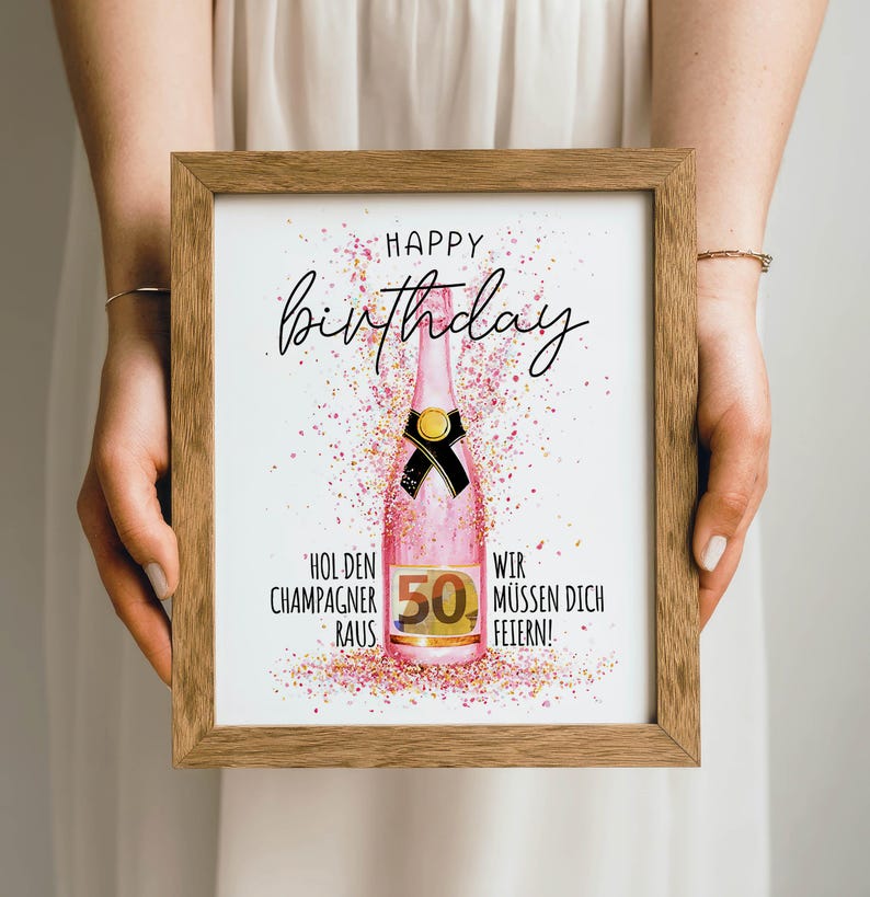 Puede incluir: Impresi&oacute;n enmarcada con marco de madera. La impresi&oacute;n presenta una botella de champ&aacute;n rosa con un lazo negro y el n&uacute;mero 50. Las palabras "Happy Birthday" est&aacute;n escritas encima de la botella, con texto alem&aacute;n debajo.