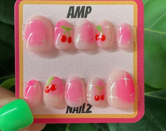 May include: A set of pink and white press-on nails with a cherry design. The nails are on a pink and yellow card with the text "AMP NAILZ".