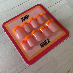 Pu&ograve; includere: Un set di dieci unghie finte rosa con un design a punta francese bicolore arancione e rosa. Le unghie sono esposte su una carta rosa e gialla con il testo "AMP NAILZ".