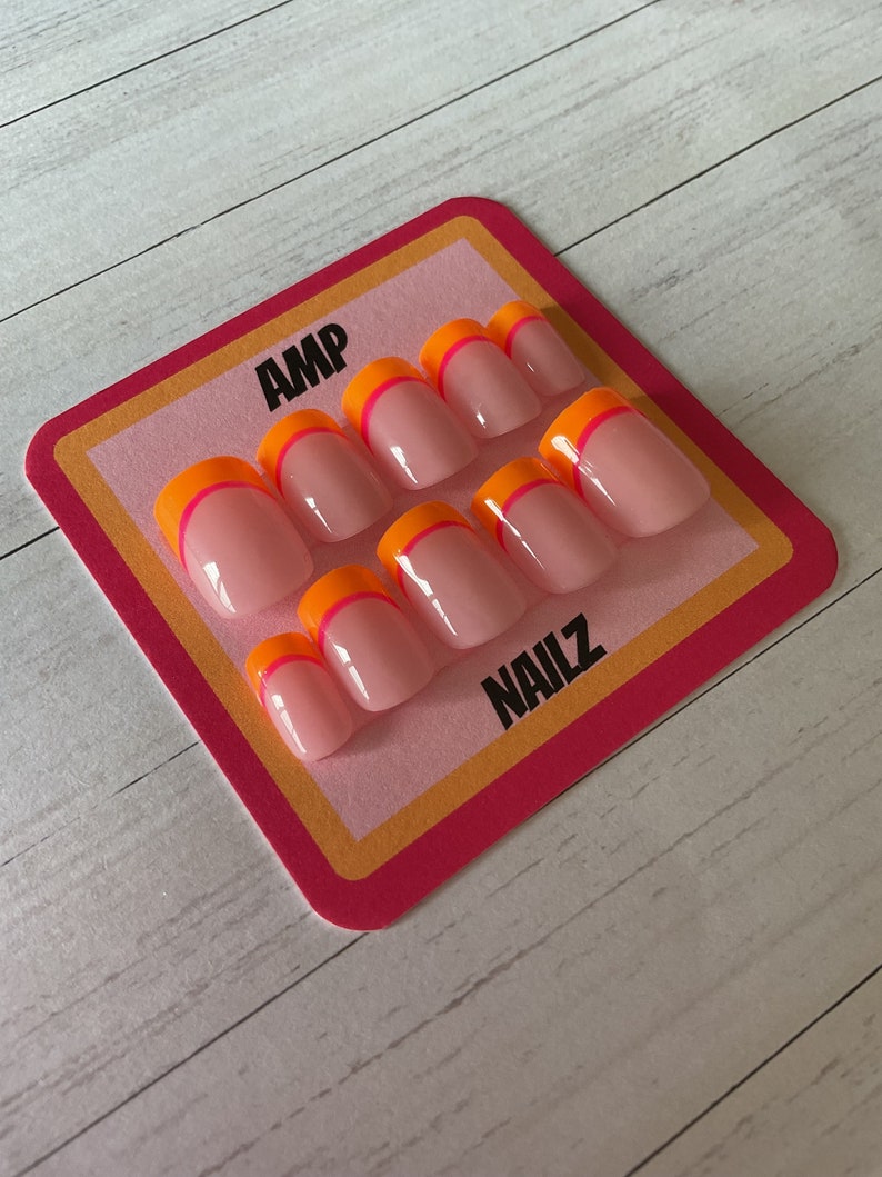 Pu&ograve; includere: Un set di dieci unghie finte rosa con un design a punta francese bicolore arancione e rosa. Le unghie sono esposte su una carta rosa e gialla con il testo "AMP NAILZ".