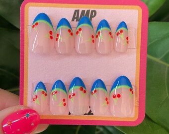 May include: A set of ten false nails with a cherry design. The nails are oval shaped and have a blue and green accent at the tip. The nails are displayed on a pink card with the word "AMP" printed on it.