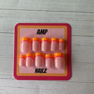 Pu&ograve; includere: Un set di 10 unghie finte rosa con un accento arancione e rosa a doppia linea sul bordo libero. Le unghie sono esposte su una carta rosa e arancione con il testo "AMP NAILZ".