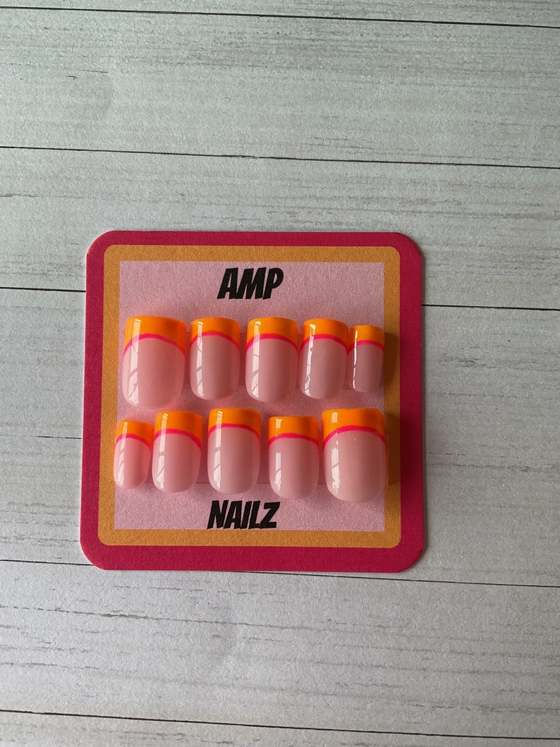 Pu&ograve; includere: Un set di 10 unghie finte rosa con un accento arancione e rosa a doppia linea sul bordo libero. Le unghie sono esposte su una carta rosa e arancione con il testo "AMP NAILZ".
