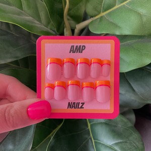Pu&ograve; includere: Un set di 10 unghie finte rosa con una doppia linea di accento arancione e rosa. Le unghie sono esposte su una carta rosa e arancione con il testo "AMP NAILZ".