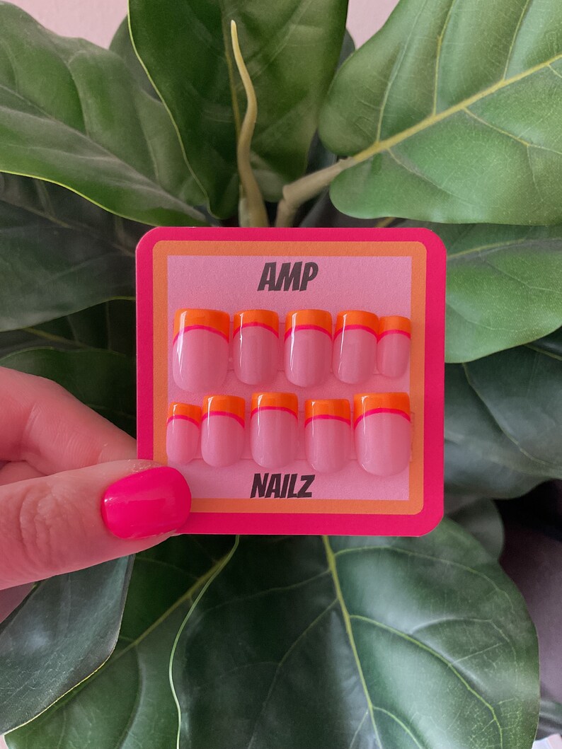 Pu&ograve; includere: Un set di 10 unghie finte rosa con una doppia linea di accento arancione e rosa. Le unghie sono esposte su una carta rosa e arancione con il testo "AMP NAILZ".