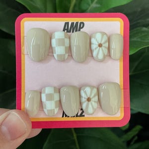 May include: A set of ten press-on nails in a variety of designs. The nails are a neutral beige color, with some featuring a white checkerboard pattern and others a white daisy design. The card has the text "AMP".