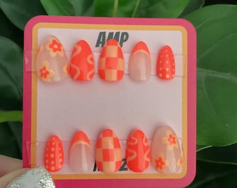 May include: A set of ten press-on nails with different designs. The nails are oval shaped and have a light pink base. The designs include flowers, checkered patterns, swirls, and polka dots. The nails are arranged in two rows on a pink and white card with the word "AMP" printed on it.