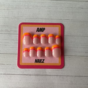 Pu&ograve; includere: Un set di dieci unghie finte con base rosa e punta doppia arancione e rosa. Le unghie sono esposte su una carta rosa e gialla con il testo "AMP NAILZ".