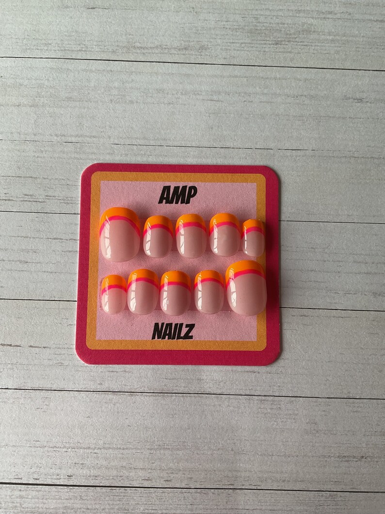 Pu&ograve; includere: Un set di dieci unghie finte con base rosa e punta doppia arancione e rosa. Le unghie sono esposte su una carta rosa e gialla con il testo "AMP NAILZ".