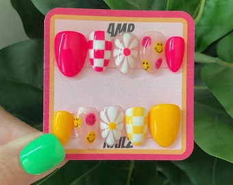 May include: A set of ten press-on nails with a pink and yellow base. The nails feature a variety of designs, including checkered, smiley faces, and daisies. The nails are displayed on a pink card with the text "4MP NAILS".