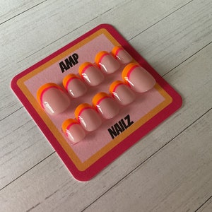 Pu&ograve; includere: Un set di dieci unghie finte con base rosa e un design a doppia linea in arancione e rosa. Le unghie sono esposte su una carta rosa e arancione con il testo "AMP NAILZ".