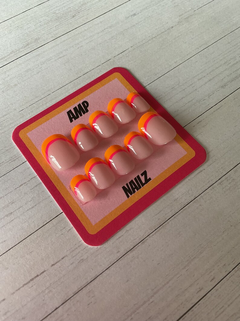 Pu&ograve; includere: Un set di dieci unghie finte con base rosa e un design a doppia linea in arancione e rosa. Le unghie sono esposte su una carta rosa e arancione con il testo "AMP NAILZ".