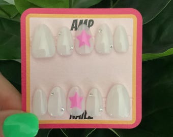 May include: A set of ten white press-on nails with pink star accents. The nails are displayed on a pink and yellow card with the text "AMP NAILS".