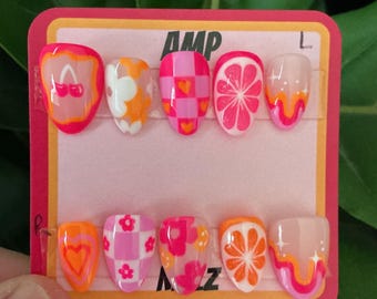 May include: A set of ten press-on nails with colorful designs. The nails feature various patterns, including hearts, flowers, and citrus fruit slices, in shades of pink, orange, and white. The card reads "AMP" in bold, black letters.