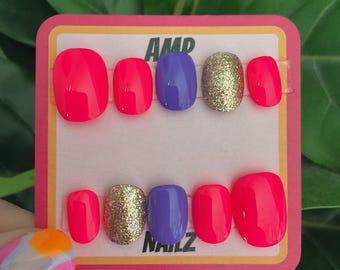Puede incluir: Un conjunto de diez uñas postizas con una variedad de colores y acabados. Las uñas están dispuestas en dos filas de cinco. La primera fila presenta dos uñas de color rosa brillante, una uña morada y dos uñas con purpurina dorada. La segunda fila presenta dos uñas de color rosa brillante, una uña con purpurina dorada y dos uñas moradas. Las uñas están etiquetadas como "AMP NAILZ".
