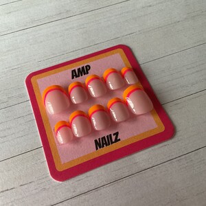 Pu&ograve; includere: Un set di dieci unghie finte con base rosa e un accento arancione e rosa a doppia linea. Le unghie sono esposte su una carta rosa e arancione con il testo "AMP NAILZ".