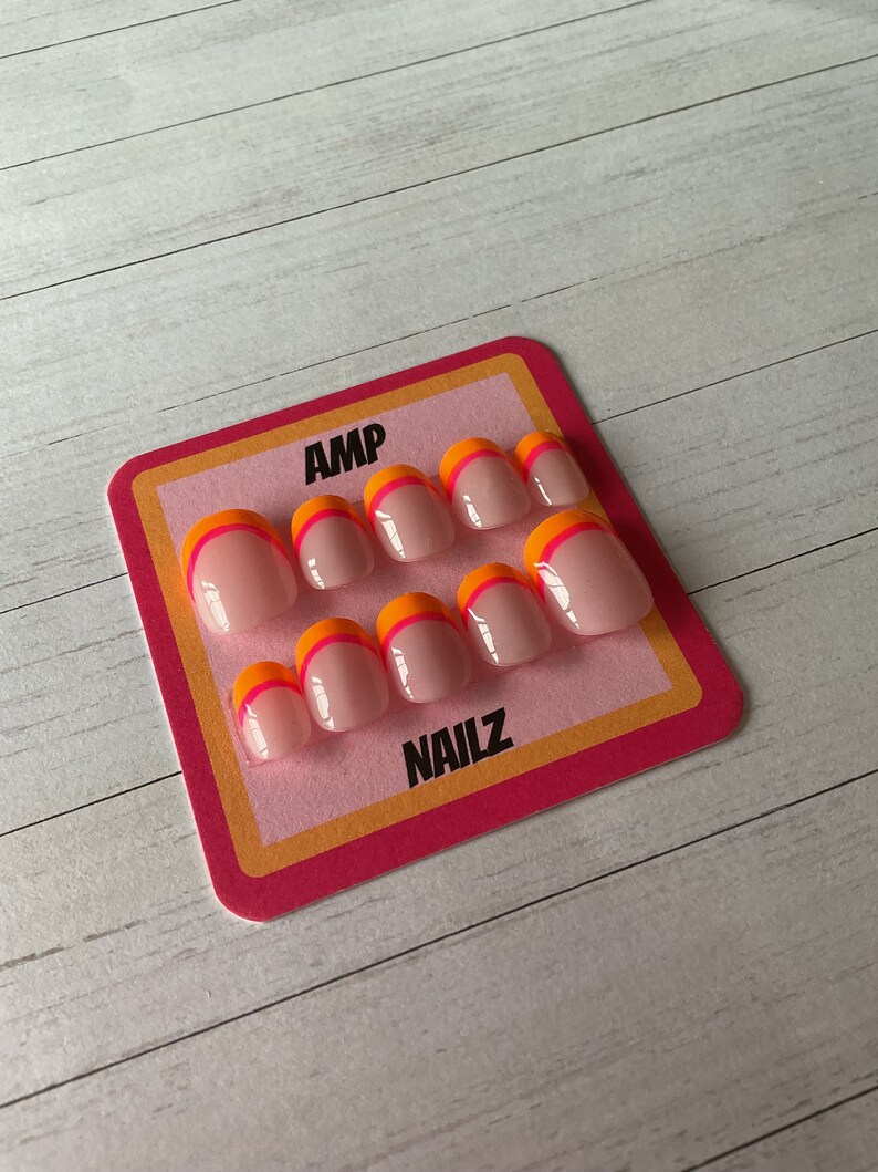 Pu&ograve; includere: Un set di dieci unghie finte con base rosa e un accento arancione e rosa a doppia linea. Le unghie sono esposte su una carta rosa e arancione con il testo "AMP NAILZ".