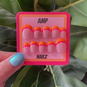 Pu&ograve; includere: Un set di dieci unghie finte rosa con un design bicolore arancione e rosa. Le unghie sono esposte su una carta rosa e arancione con il testo "AMP NAILZ".